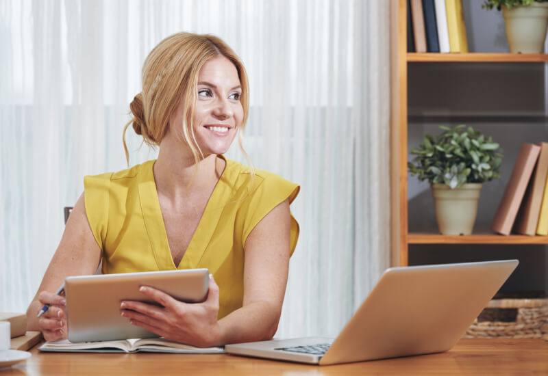 young woman in yellow blouse smiling holding ipad at desk next to laptop