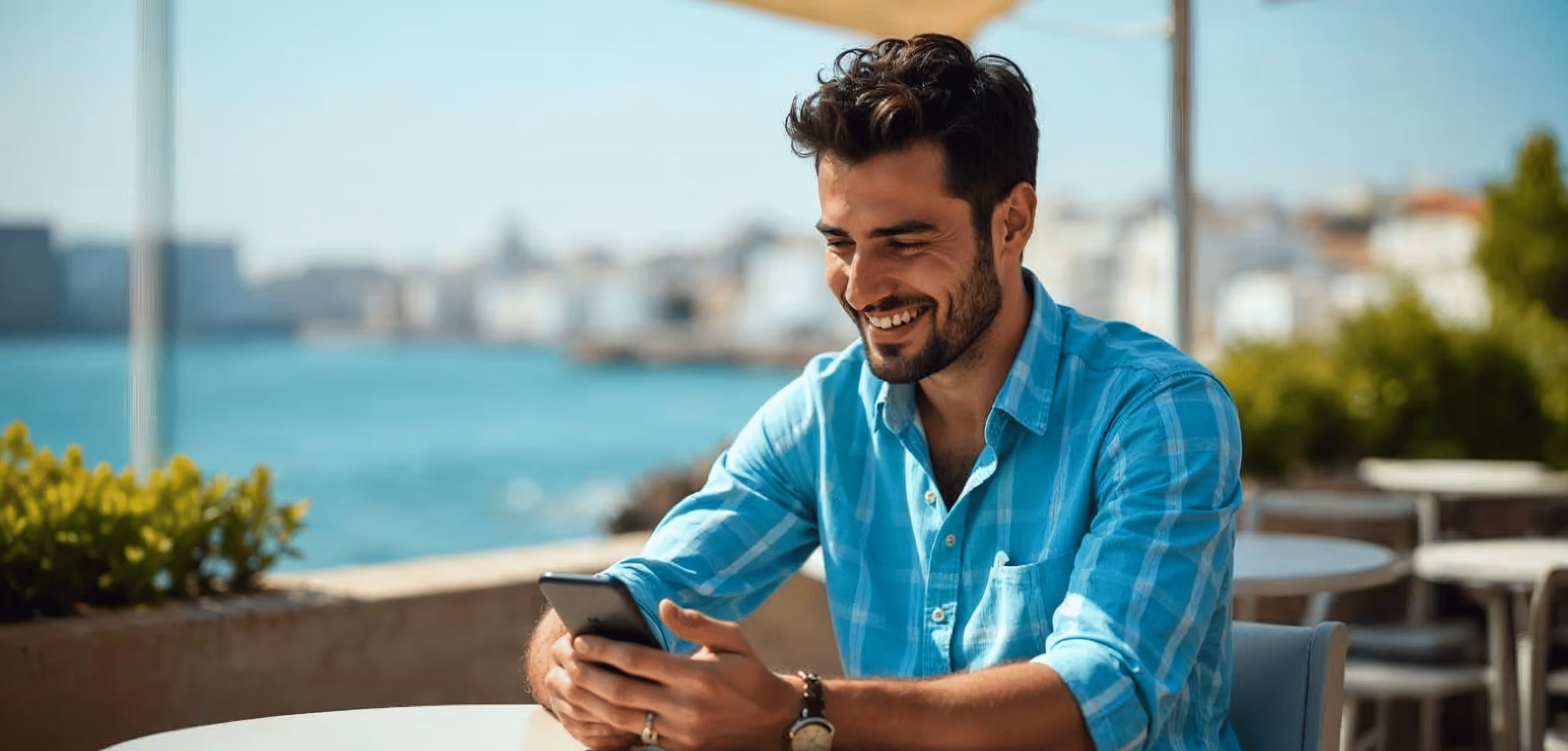 Smiling man in a blue plaid shirt sitting outdoors at a table, looking at his smartphone with a waterfront cityscape in the background.