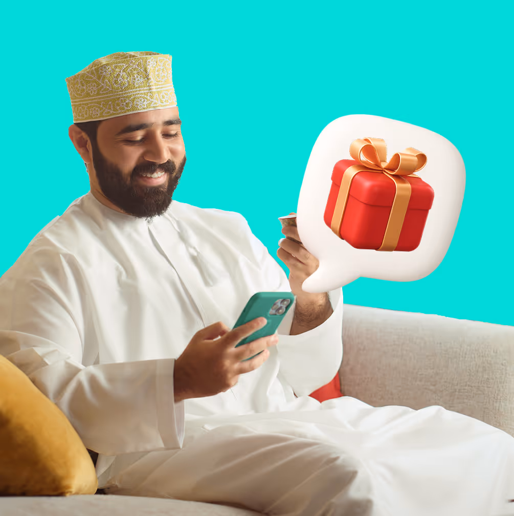رجل مبتسم يرتدي ملابس بيضاء تقليدية وقبعة منقوشة يجلس على أريكة وينظر إلى هاتفه مع فقاعة دردشة تظهر صندوق هدايا أحمر ملفوف بشريط ذهبي.