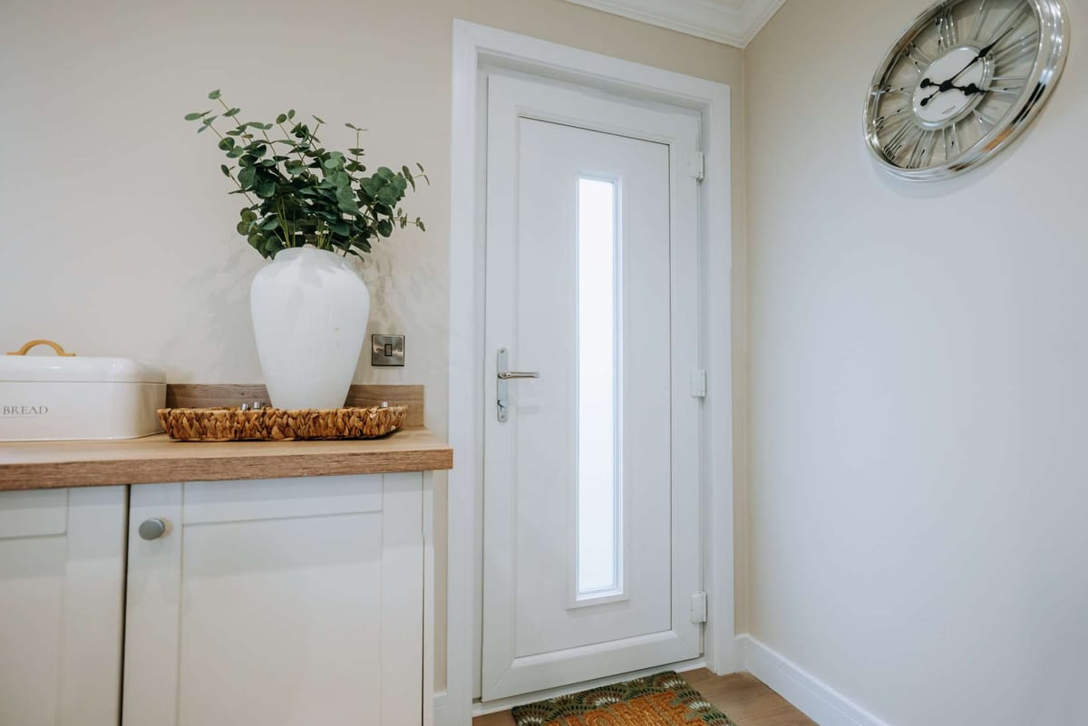 Bright Kitchen Corner with White Door, Large Clock, and Decorative Elements Including a Tall Vase with Greenery, Bread Box, and Woven Basket on Wooden Countertop