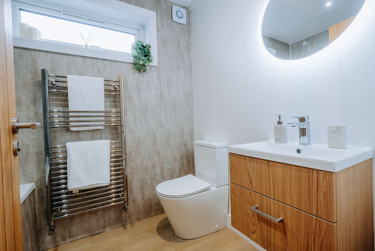 Modern Bathroom with Wood Accents Featuring a Minimalist Vanity and Backlit Mirror, Chrome Heated Towel Rail with White Towels, Compact Toilet, and Decorative Greenery Under a Small Window