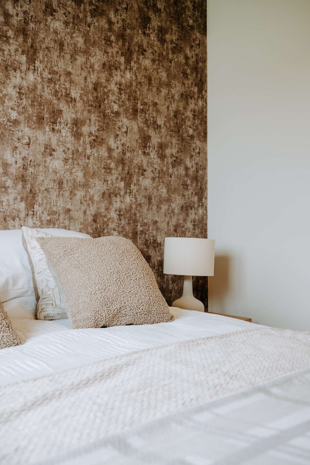Cozy Bedroom with Textured Brown Accent Wall, Soft Beige Pillows, White Bedding, and Minimalist Table Lamp
