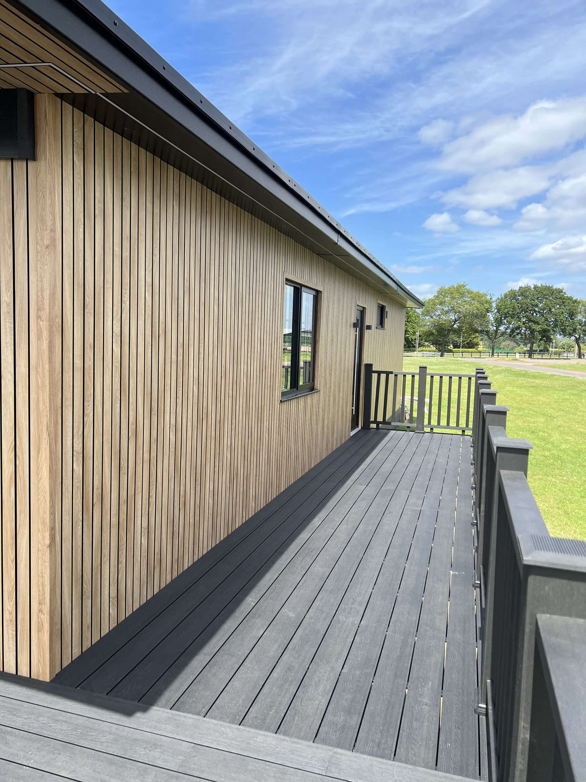 Modern Wooden Siding House with Black Trim and Spacious Grey Deck Overlooking Expansive Green Lawn and Blue Sky with Light Clouds