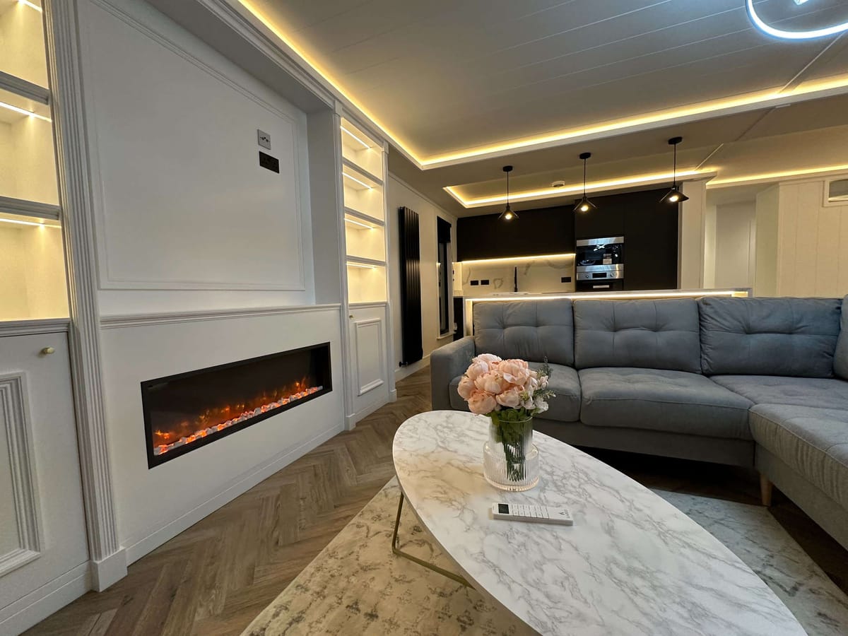 Modern Living Room with Cozy Electric Fireplace, Elegant Gray Sectional Sofa, Marble Coffee Table with Floral Centerpiece, and Sleek Open Kitchen Featuring Pendant Lighting and Built-In Appliances