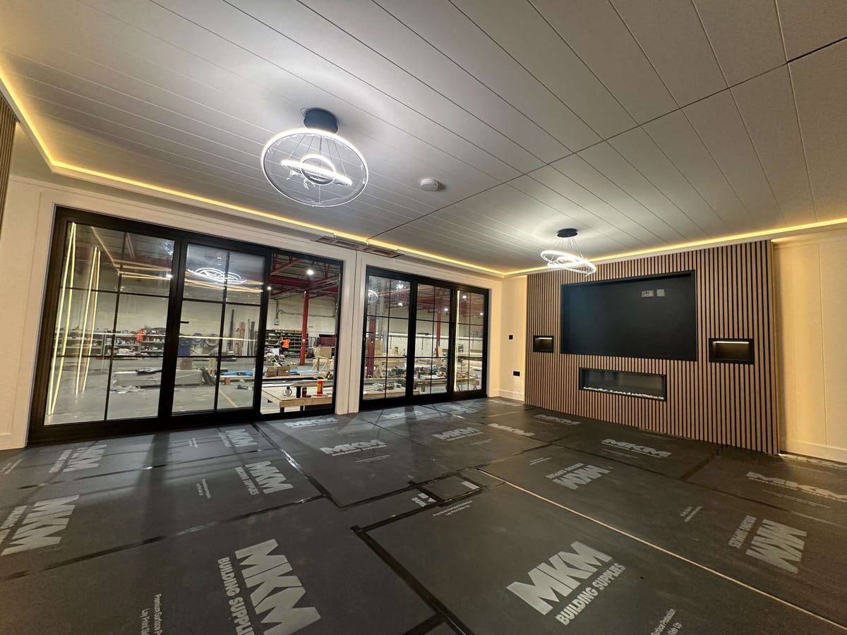 Modern Interior with Suspended Ceiling, Large Glass Doors Overlooking Workshop, and Wood-Panelled Wall Featuring Integrated Screen and Ambient Lighting