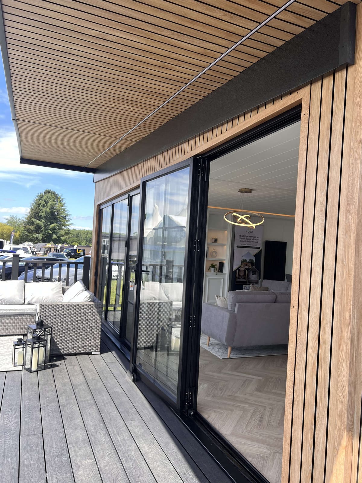 Modern Patio with Sliding Glass Doors Leading to Cozy Indoor Living Space, Featuring Wooden Cladding and Ceiling, Outdoor Wicker Furniture with Cushions, Lanterns, and Scenic View of Trees and Sky.