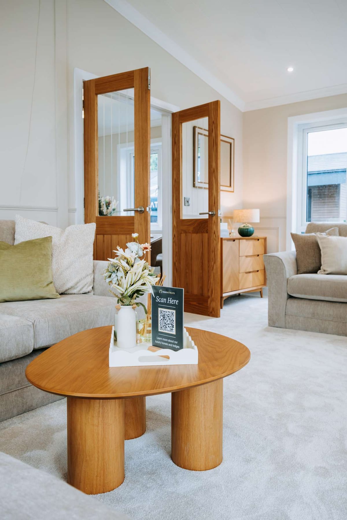 Cozy Modern Living Room with Wooden Furniture and Neutral Tones Featuring a Stylish Coffee Table with Decorative Tray and Floral Arrangement, Open Wooden Doors Leading to a Bright Interior, and Soft Plush Carpeting.
