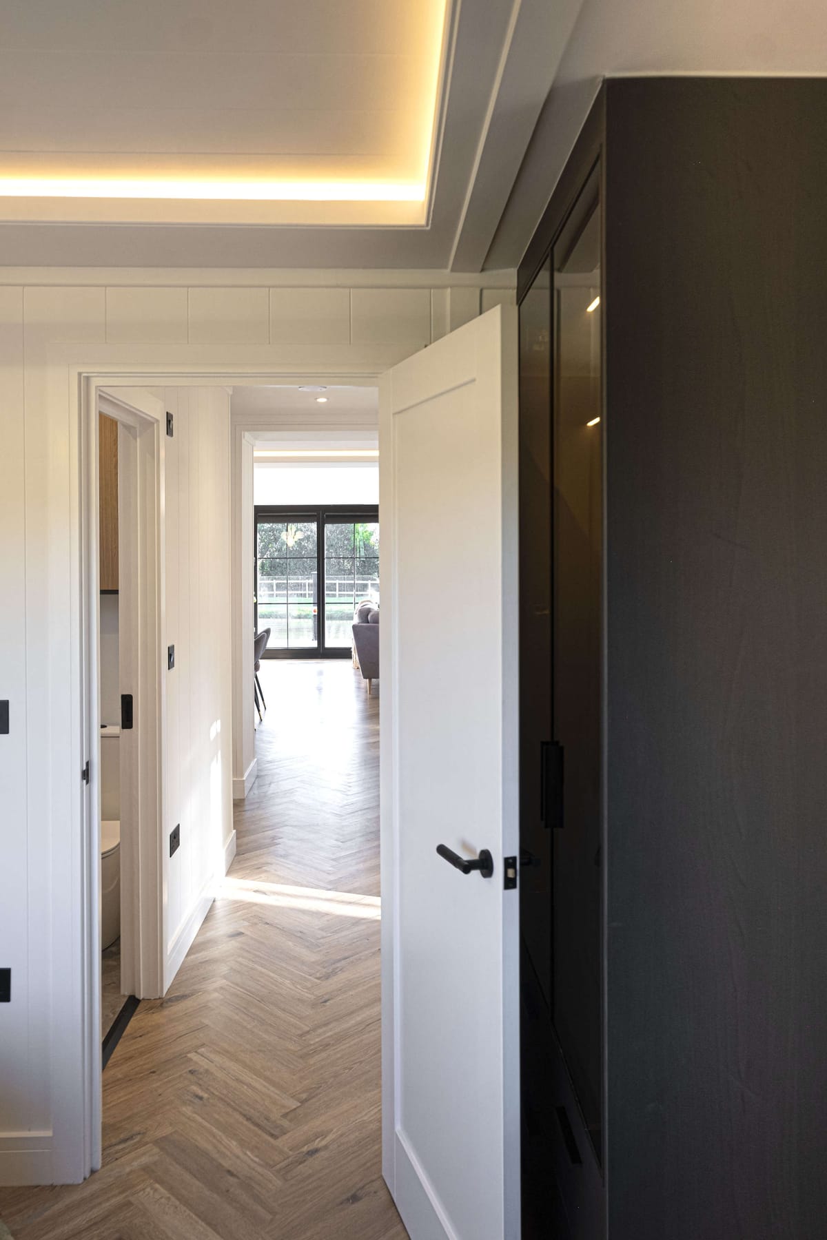 Modern Interior Hallway with Open White Door, Herringbone Wooden Flooring, and Glass Patio Doors Leading to Bright Living Room