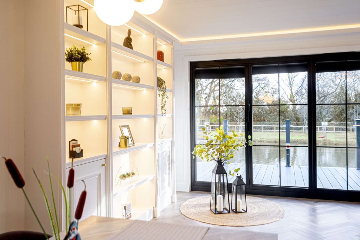 Modern Living Room with Illuminated Shelving, Large Windows Overlooking a Tranquil Water Scene, and Elegant Decorative Accents Including Lanterns and Greenery