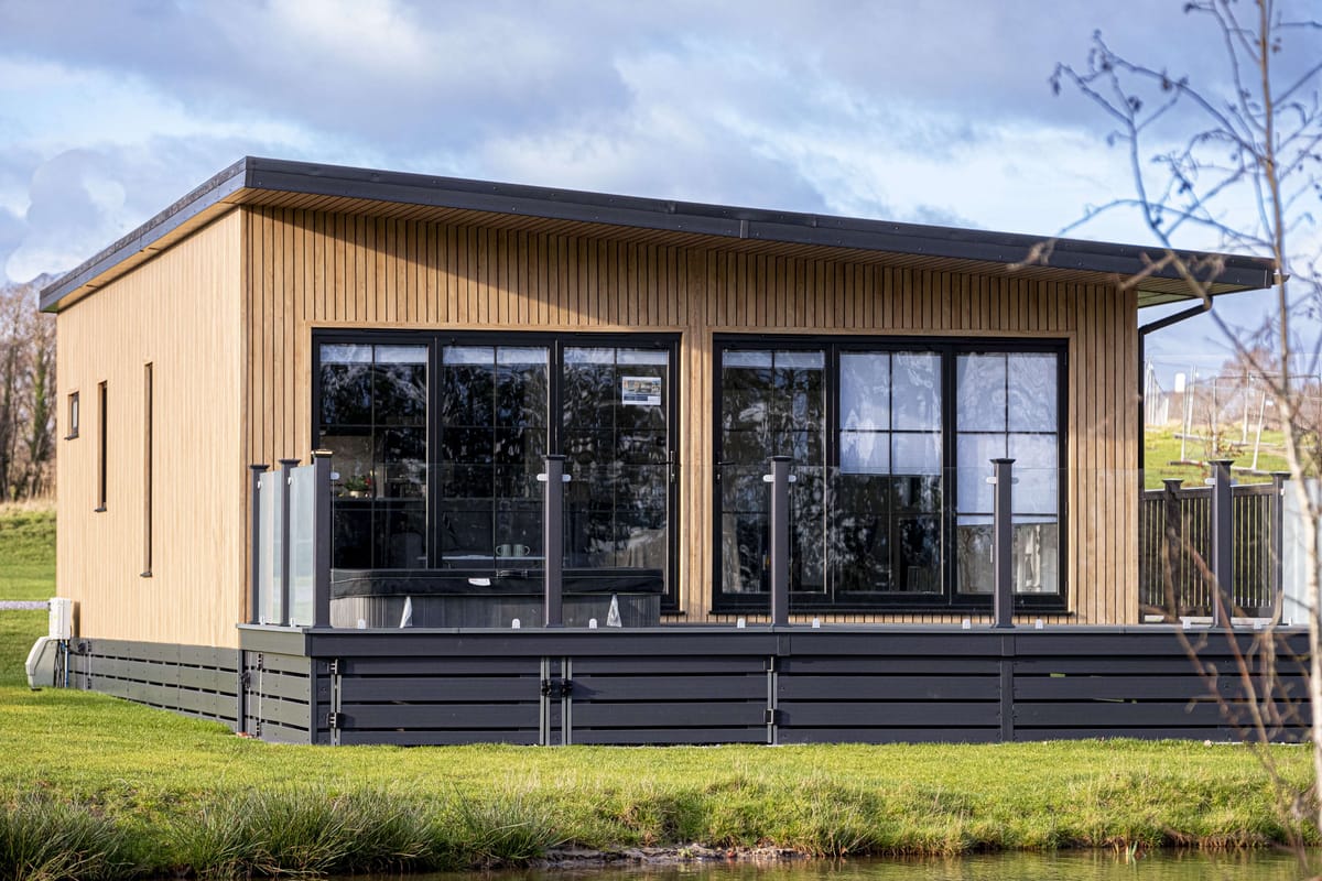 Modern Wooden Cabin with Large Glass Windows and Black Patio Fencing Set in Lush Green Countryside Under Cloudy Sky