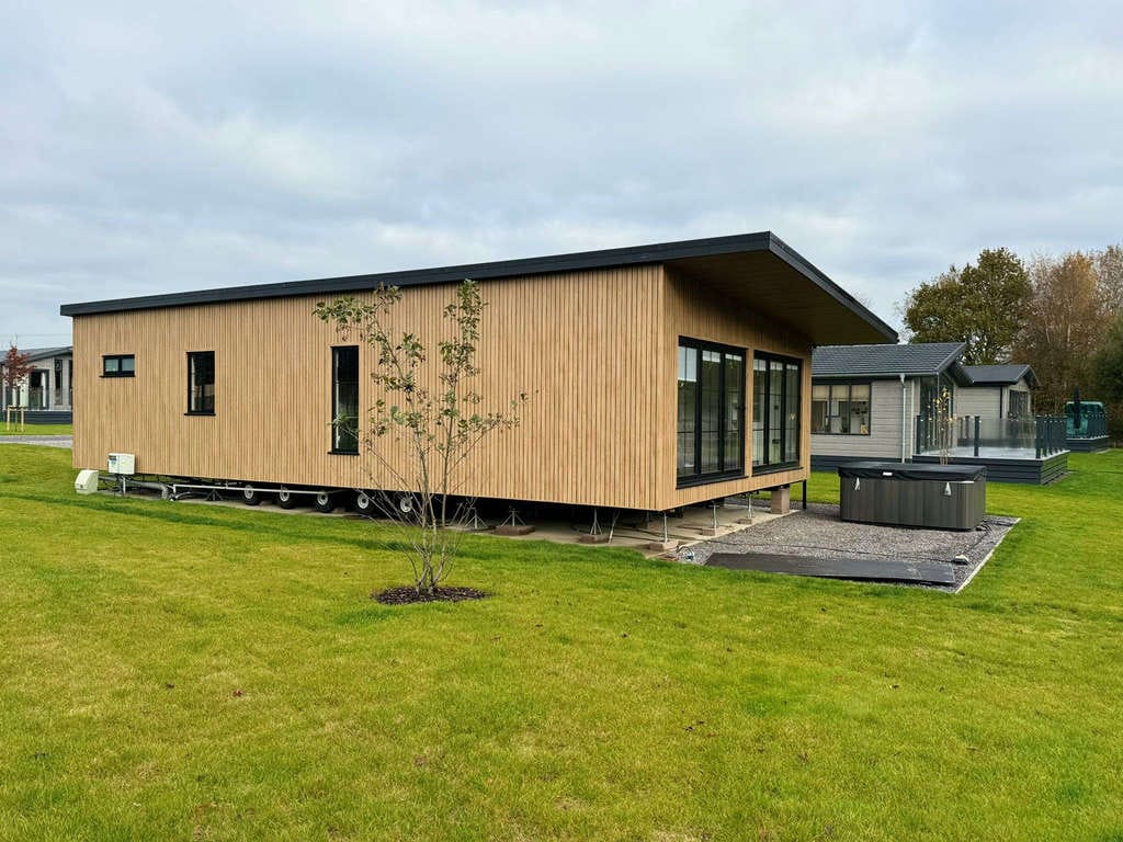 Modern Wooden Cabin with Large Windows and Sloped Roof on Expansive Green Lawn with Small Tree and Outdoor Hot Tub in Scenic Countryside Setting