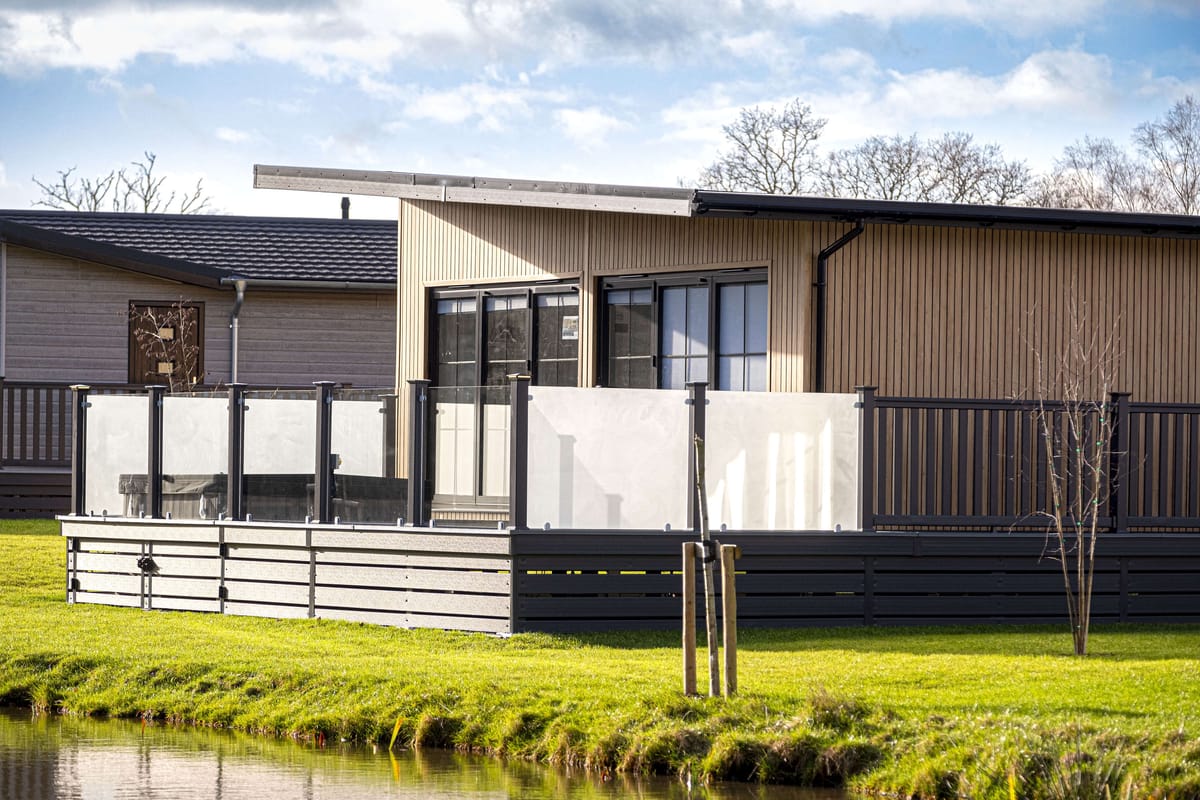 Modern Wooden Cabin with Glass-Enclosed Deck Overlooking Serene Pond and Lush Green Lawn under a Partly Cloudy Sky