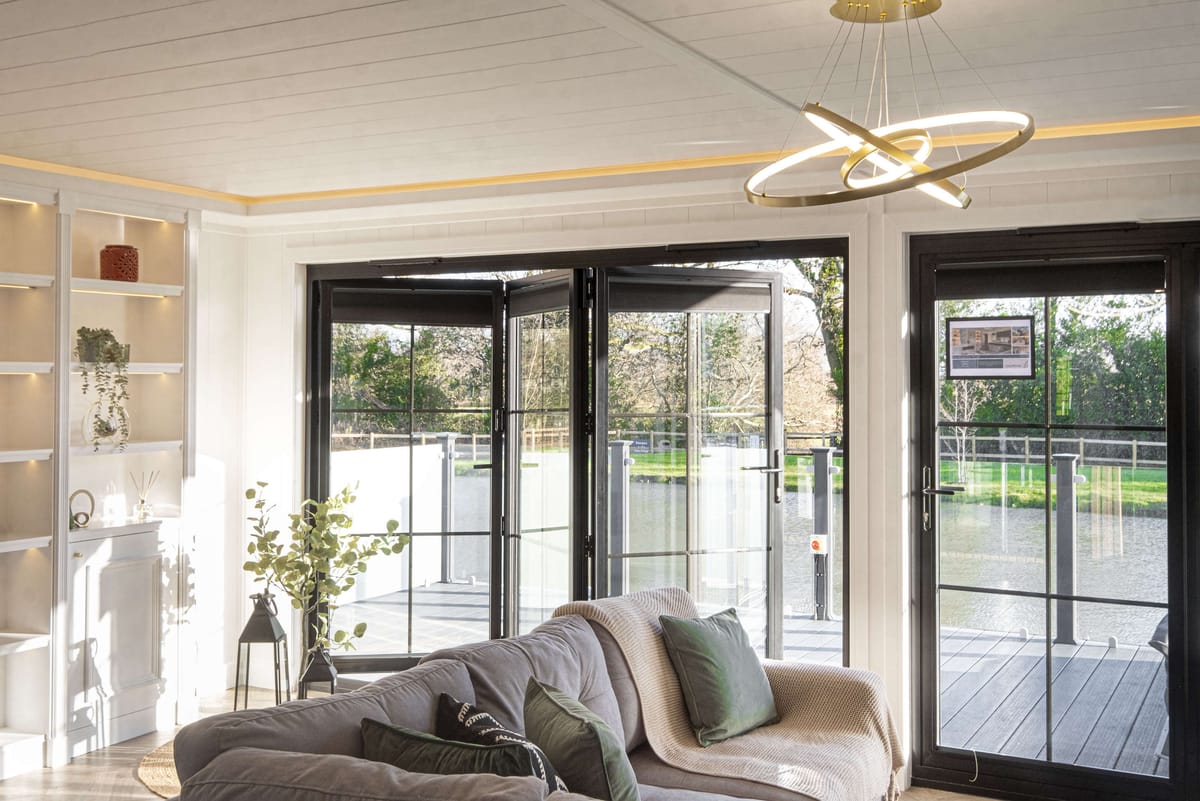 Modern Living Room with Large Glass Doors Opening to Scenic Lakeside View, Elegant Minimalist Decor, Built-In Shelving with Plants, Cozy Gray Sofa with Green Cushions, and Contemporary Circular Chandelier.