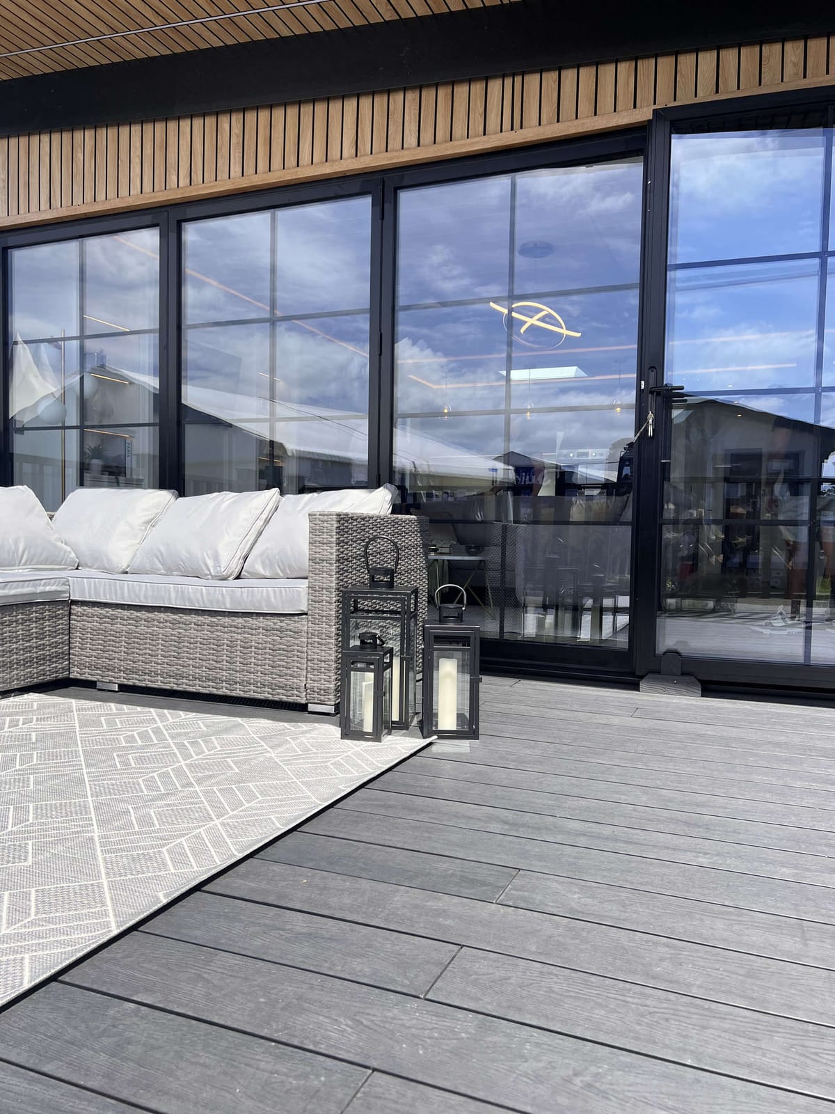 Modern Outdoor Patio with Wicker Sofa, Geometric Rug, and Decorative Lanterns Against Large Glass Doors Reflecting Blue Sky and Cloud Patterns
