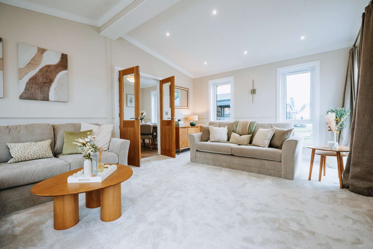 Elegant Contemporary Living Room with Neutral Tones Featuring Two Comfortable Sofas, Abstract Wall Art, Wooden Coffee Table, and Abundant Natural Light Through Large Windows and Open Wooden Doors Leading to Dining Area