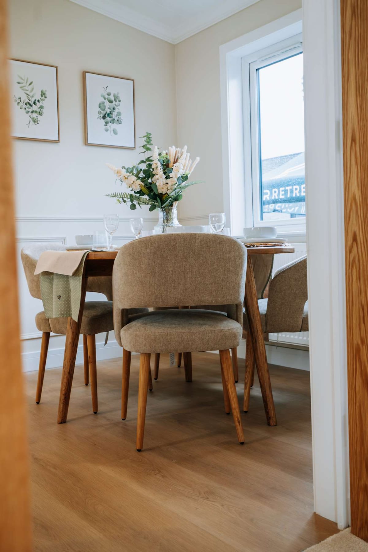 Cozy Dining Area with Elegant Wooden Furniture, Warm Beige Upholstered Chairs, Floral Centerpiece, and Botanical Wall Art in a Bright Room with Wooden Flooring and Large Window