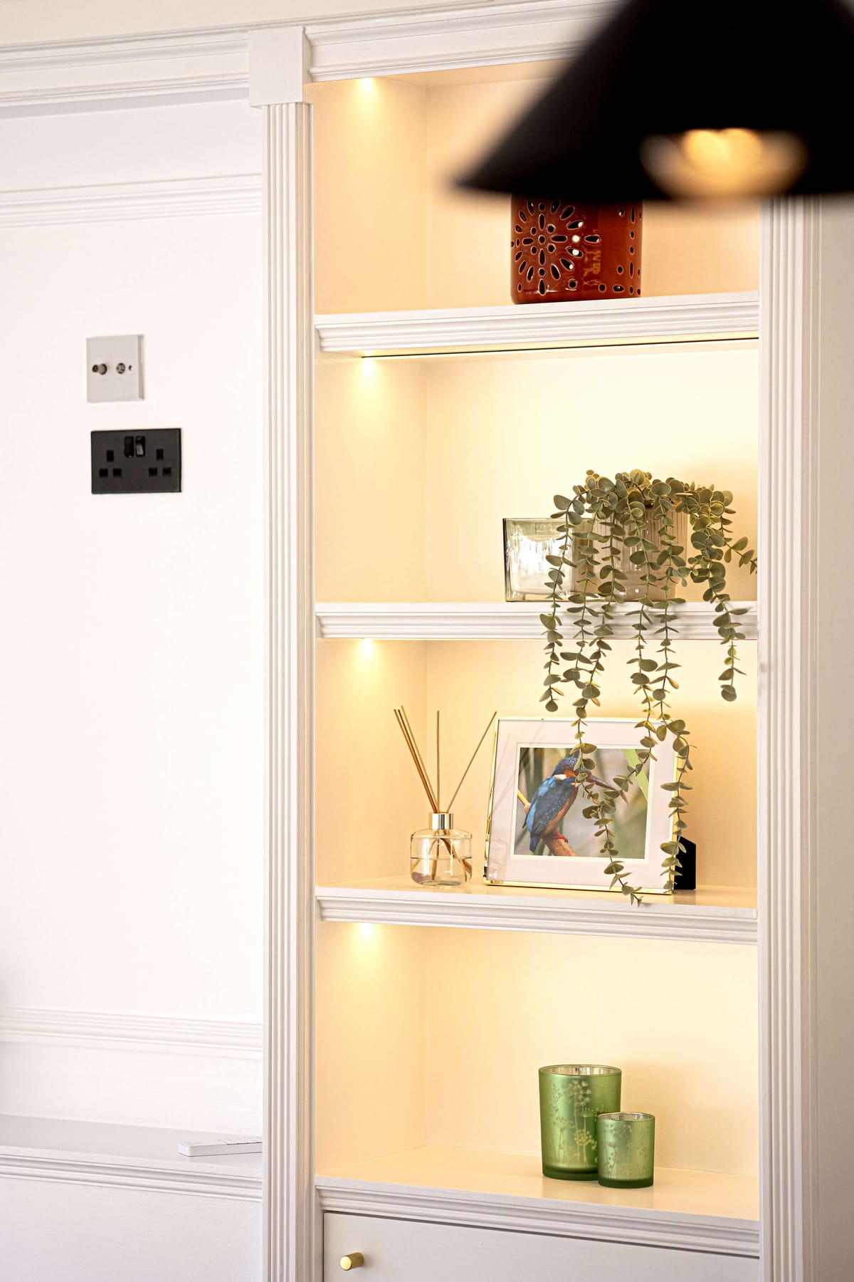 Illuminated White Shelving Unit with Decorative Items Including Red Vase, Framed Bird Photo, Glass Reed Diffuser, Green Candles, and Hanging Plant in Cozy Interior Setting