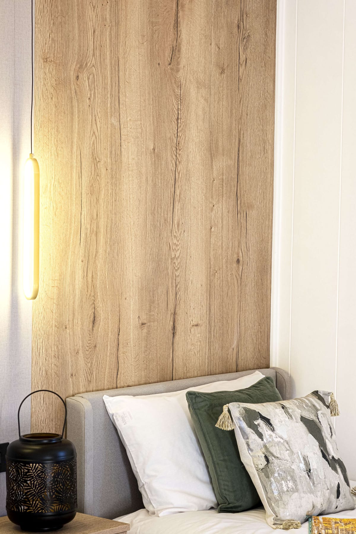 Cozy Bedroom Corner with Wooden Panel Accent Wall, Soft Cushions, Modern Hanging Light, and Decorative Black Lantern on Bedside Table