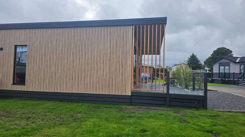 Modern Wooden Cabin with Vertical Slat Design and Adjacent Deck in Grassy Landscape Under Overcast Sky