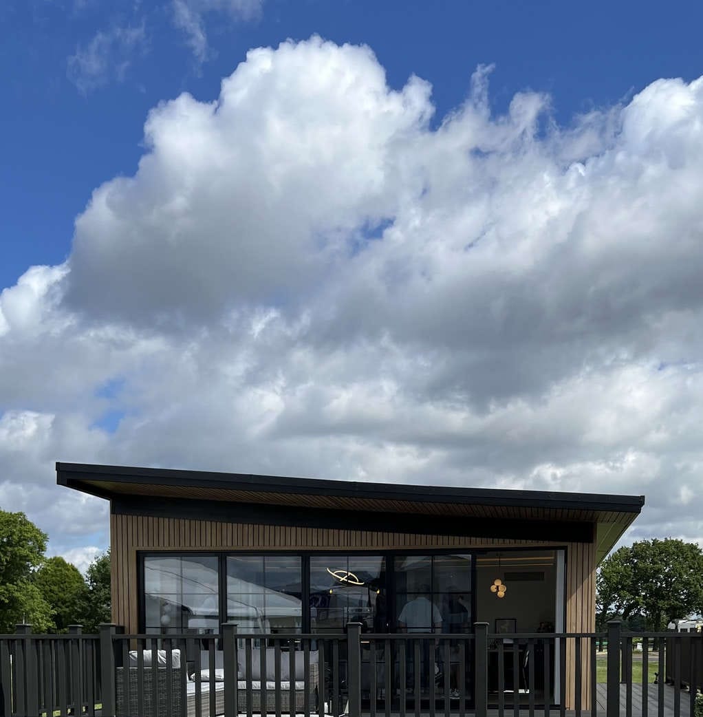 Modern Wooden Cabin with Large Glass Windows and Black Fencing Under a Bright Blue Sky with Fluffy White Clouds