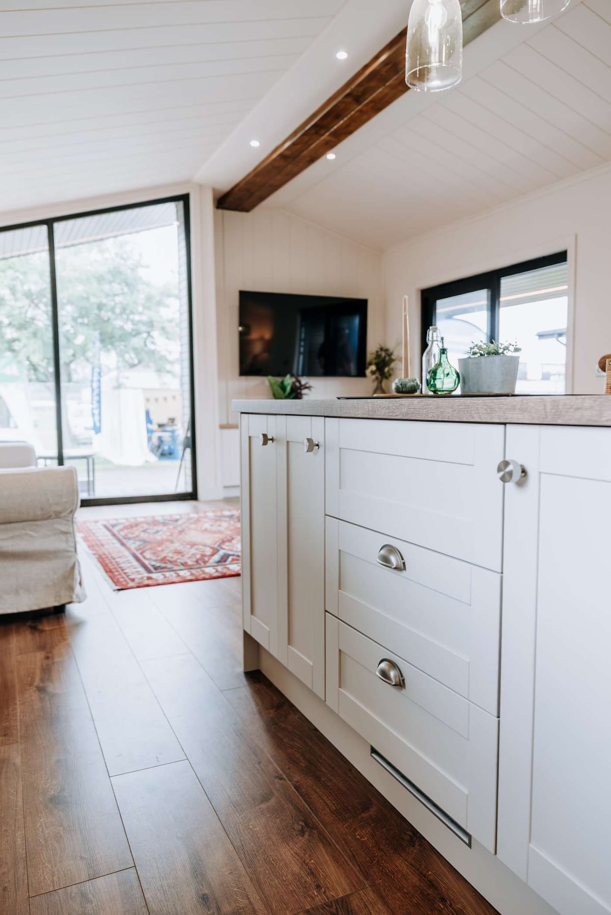 Modern Open-Concept Living Room and Kitchen with White Cabinets, Wooden Floor, and Large Windows Overlooking a Patio, Featuring Minimalist Decor and a Mounted TV