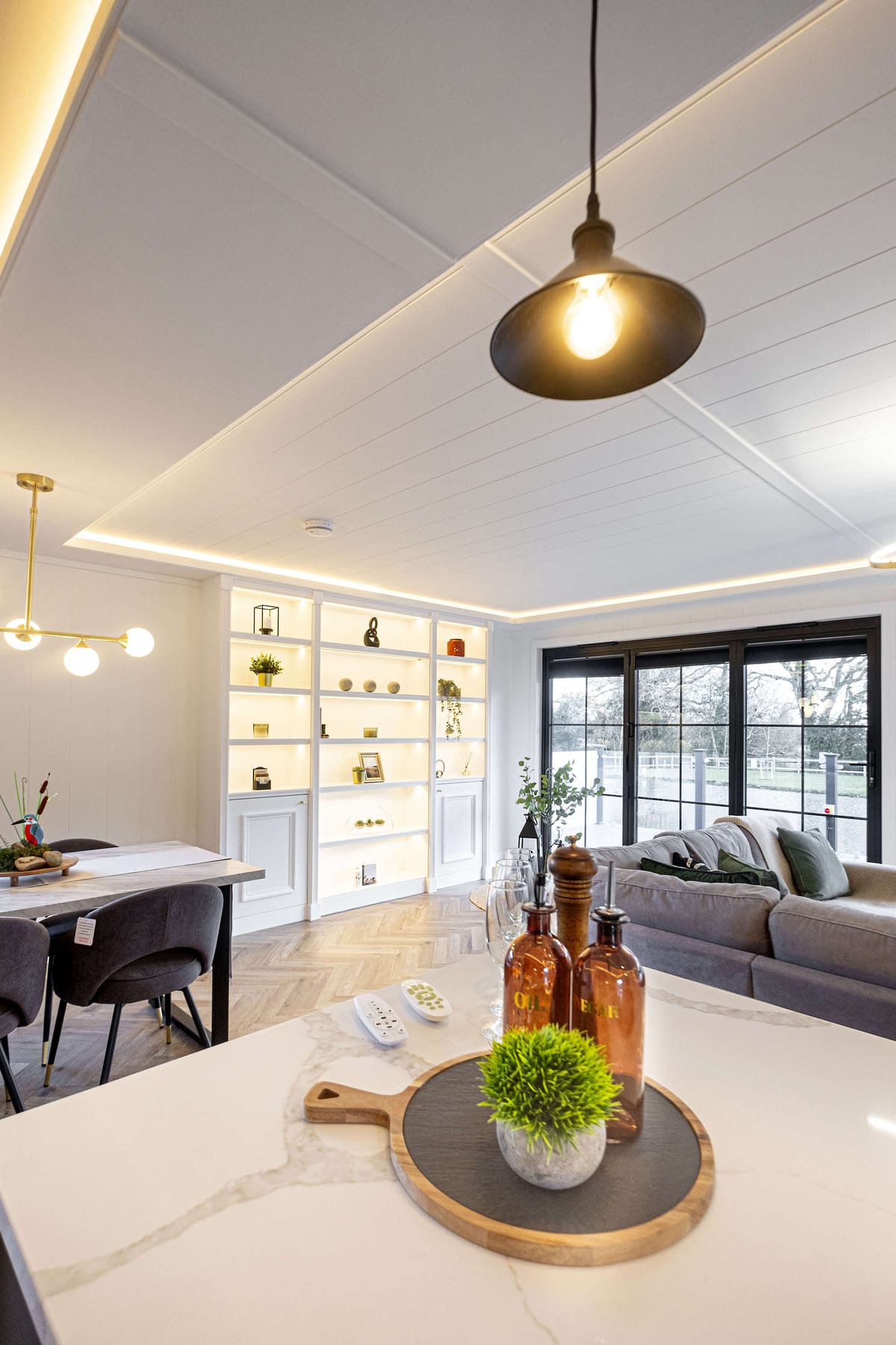 Modern Open-Concept Living Space with Elegant Lighting and Stylish Décor Featuring a Cozy Sofa, Marble Kitchen Island, and Illuminated Shelving with Decorative Accents
