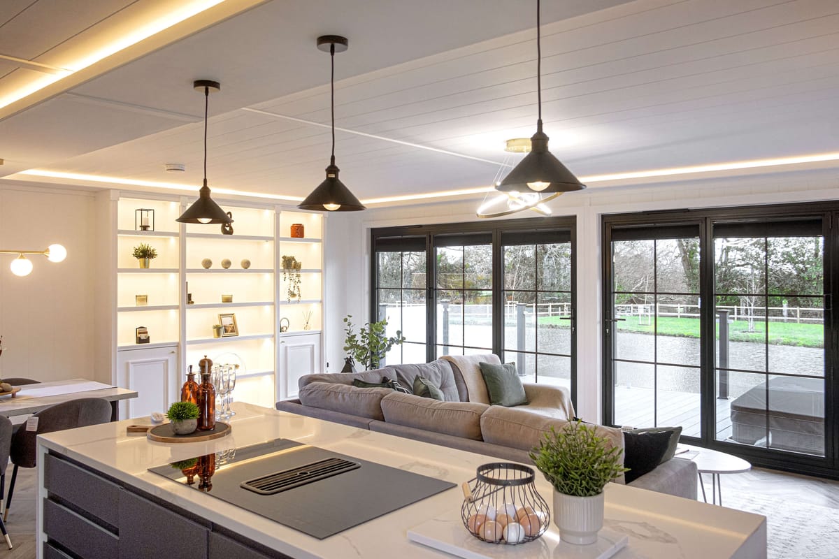 Modern Open-Plan Living Room and Kitchen with Elegant Pendant Lighting, Cozy Gray Sofa, Large Sliding Glass Doors Overlooking Scenic Backyard, and Stylish Shelving Displaying Decorative Items