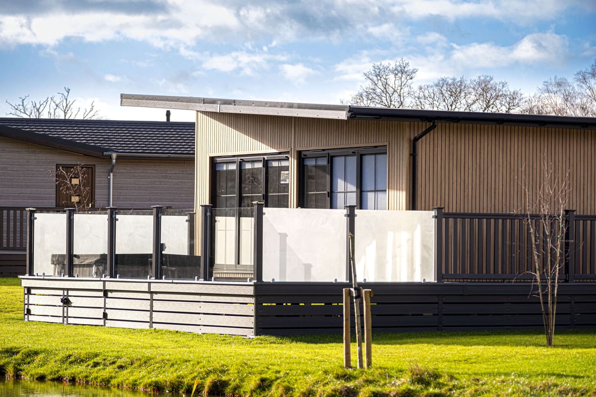 Modern Wooden House with Glass-Enclosed Deck Overlooking Lush Green Lawn and Tranquil Pond Under Blue Sky with Scattered Clouds