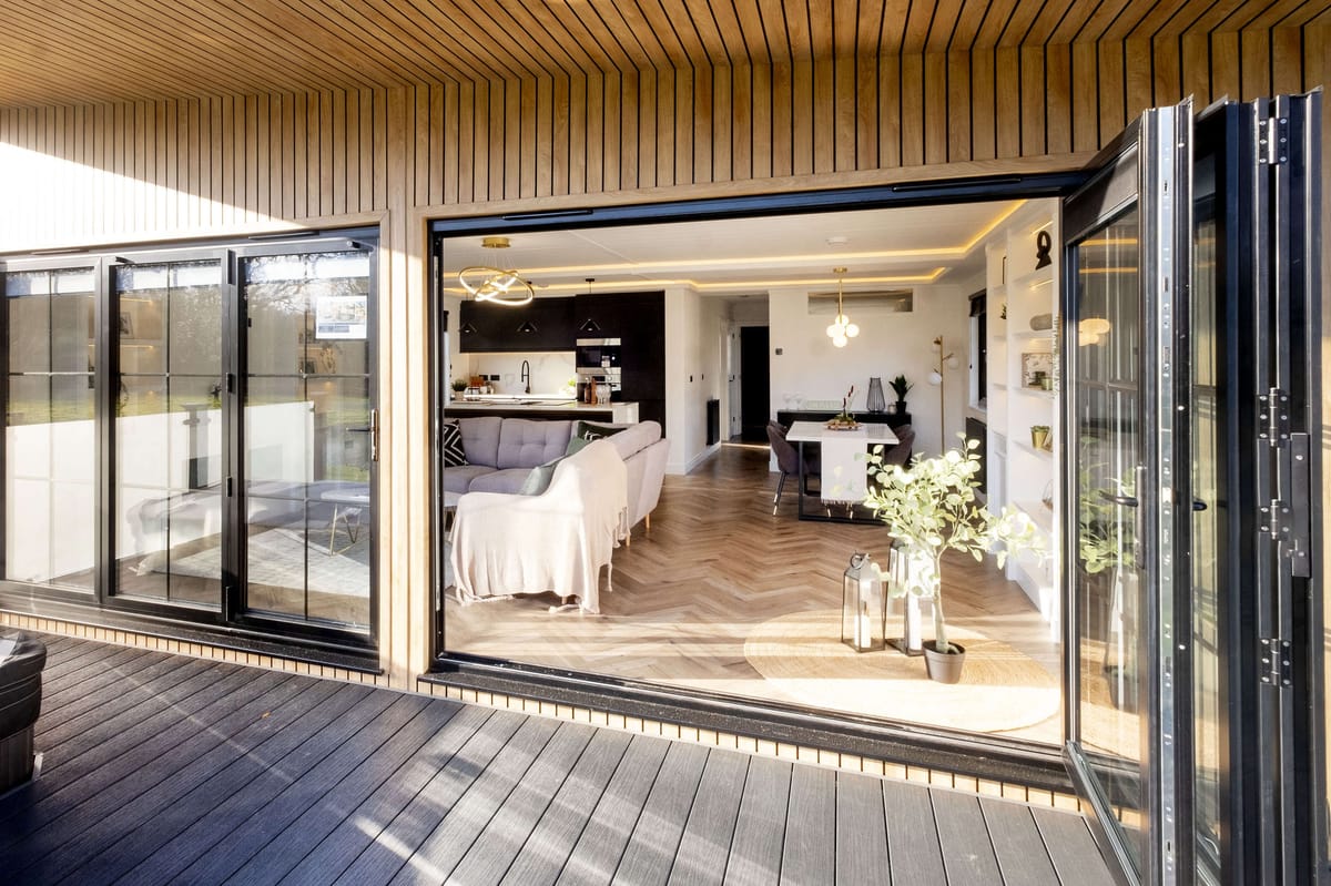 Modern Open-Plan Living Space with Wooden Interior and Large Glass Doors Leading to Outdoor Decking Area, Featuring Cozy Furniture, Stylish Lighting, and Elegant Decor Elements.