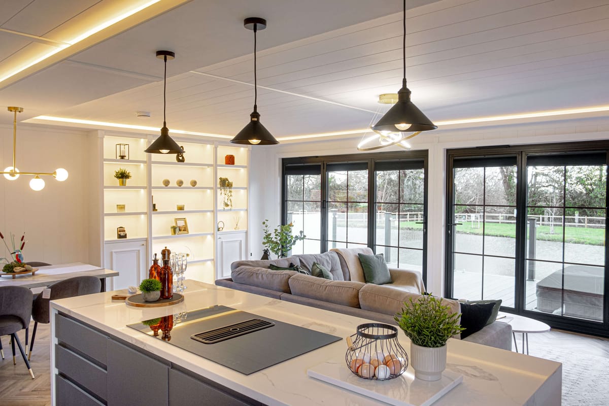Modern Open-Concept Living Space with Minimalist Kitchen Island, Cozy Sectional Sofa, Large Glass Doors Overlooking Scenic Outdoors, and Illuminated Shelving for Decorative Accents