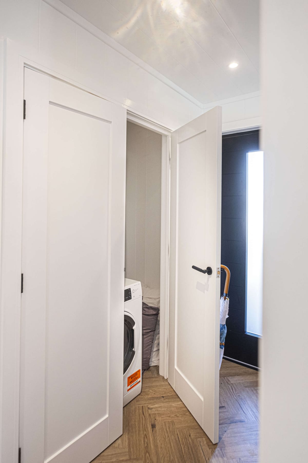Compact Laundry Nook with Modern Washing Machine Behind White Paneled Door in Bright Room with Wooden Herringbone Floor