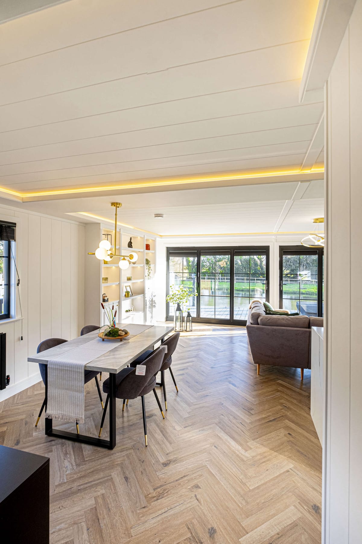 Modern Open-Plan Living and Dining Area with Herringbone Flooring, Contemporary Lighting, and Large Glass Doors Overlooking Scenic Outdoor View