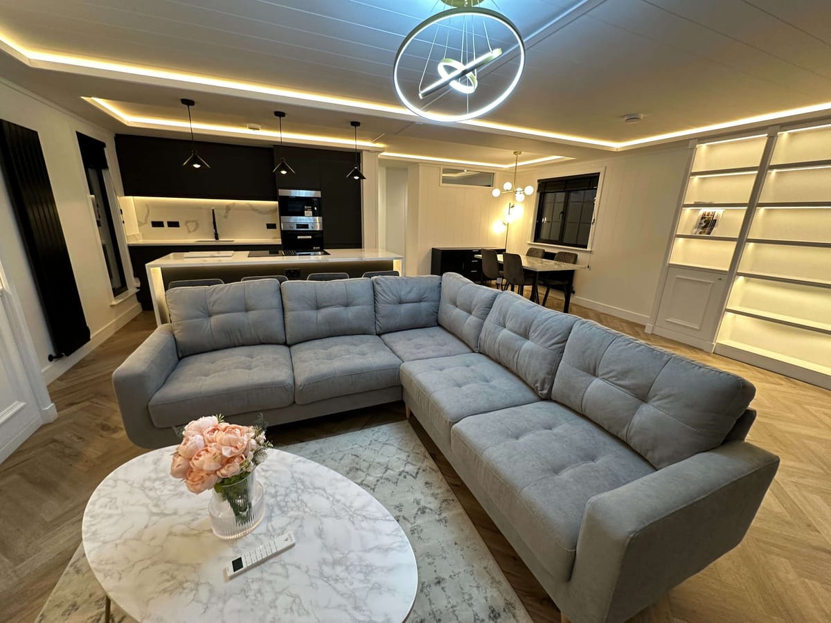 Modern Open-Concept Living Room with Gray Sectional Sofa, Marble Coffee Table, and Sleek Kitchen Featuring Black Cabinets and Pendant Lighting, Illuminated by Stylish Ceiling Fixtures and Ambient Lighting