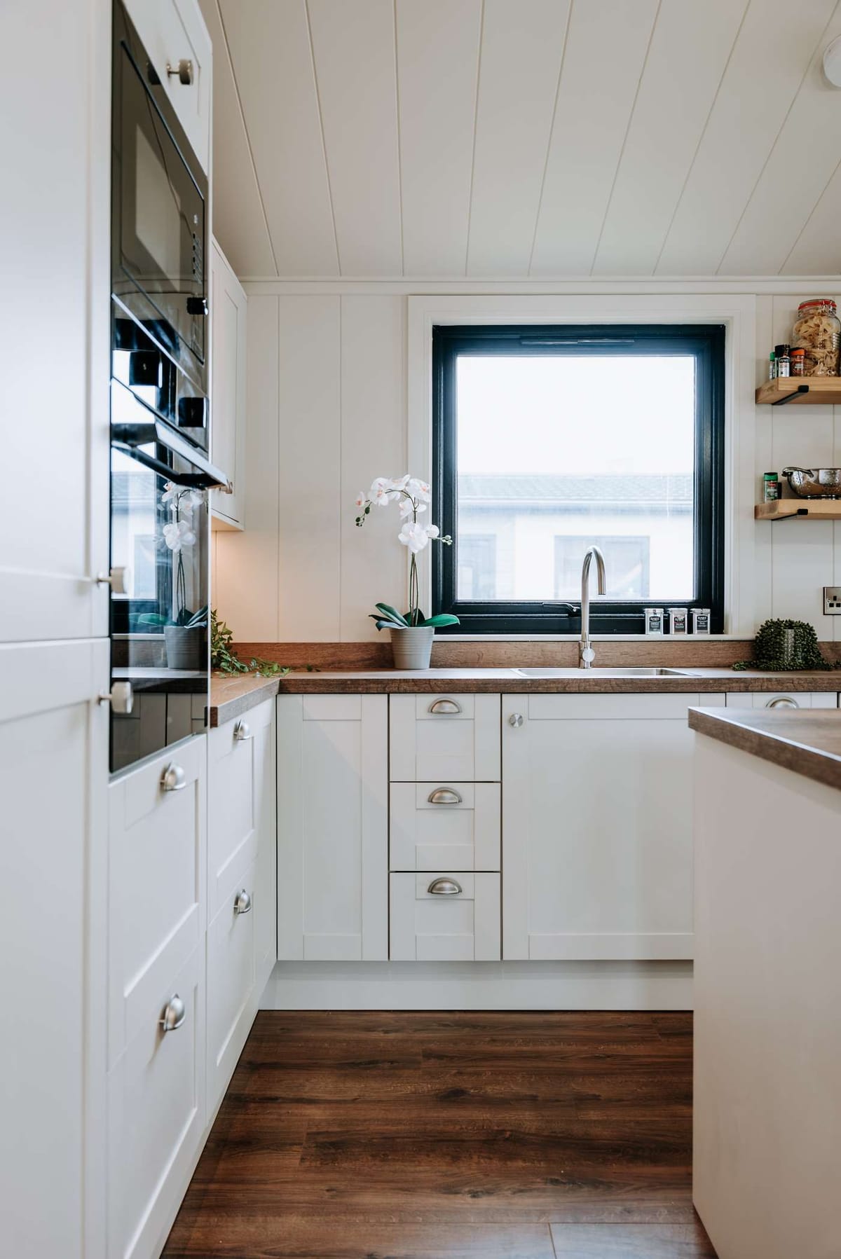Modern Kitchen Interior with White Cabinets, Wood Countertops, and Large Window Featuring Potted Orchid and Open Shelving with Assorted Jars
