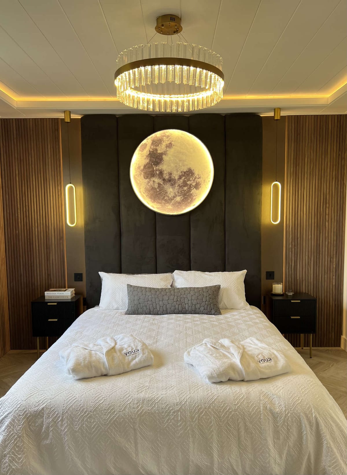 Luxurious Bedroom with Illuminated Moon Wall Art, Elegant Chandelier, and Modern Bedding Decor Featuring Soft White Linens and Cozy Robes