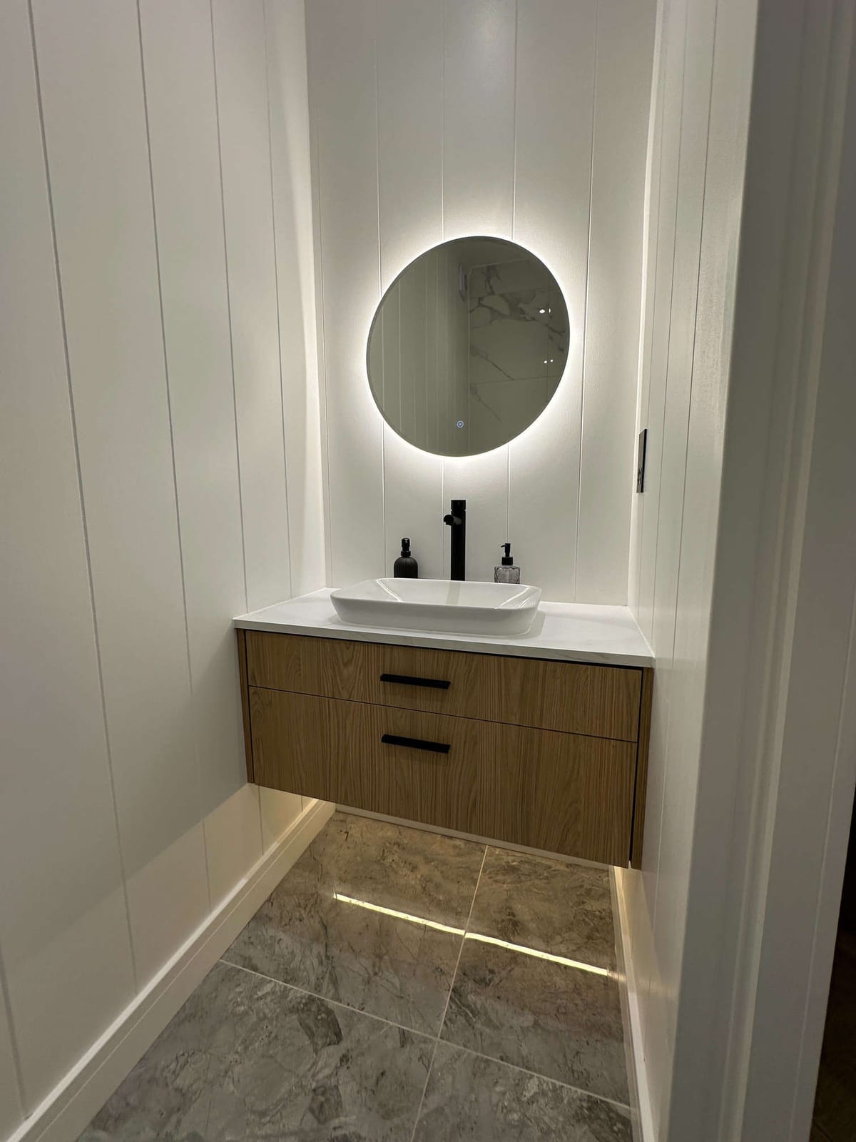 Modern Bathroom Vanity with Circular Backlit Mirror and Sleek White Basin on Wooden Floating Cabinet Against Vertical White Panel Walls and Polished Gray Marble Floor Tiles