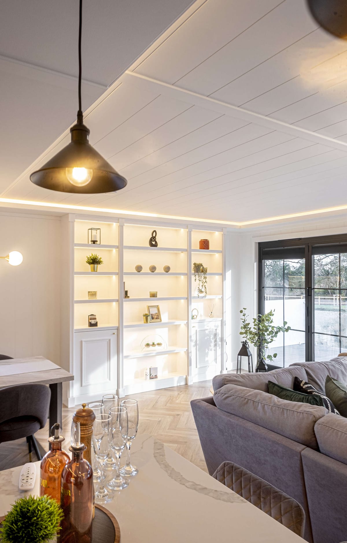 Modern Living Room with Illuminated White Shelving, Cozy Gray Sofa, and Stylish Decor Featuring a Pendant Light, Glass Doors, and Natural Light Accents
