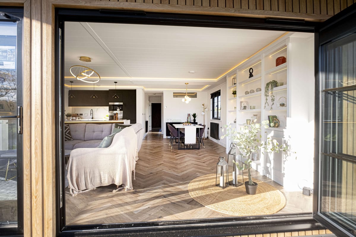 Modern Open-Plan Living Room and Kitchen with Elegant Décor, Large Windows, and Wooden Accents Featuring Cozy Seating, Dining Area, and Stylish Shelving Displaying Plants and Decorative Items.
