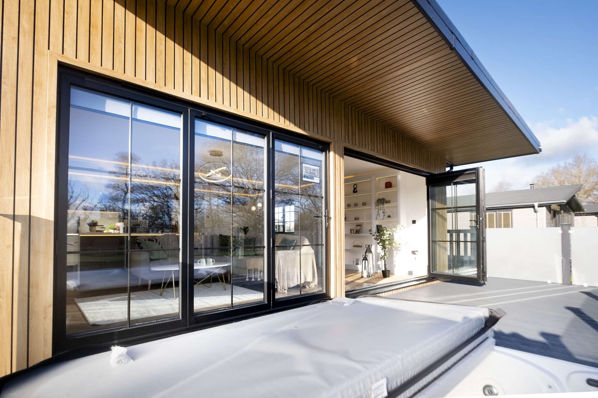 Modern Wooden Patio with Large Glass Sliding Doors Leading to a Cozy Interior Featuring Shelves and Decorative Plants, Under a Clear Blue Sky