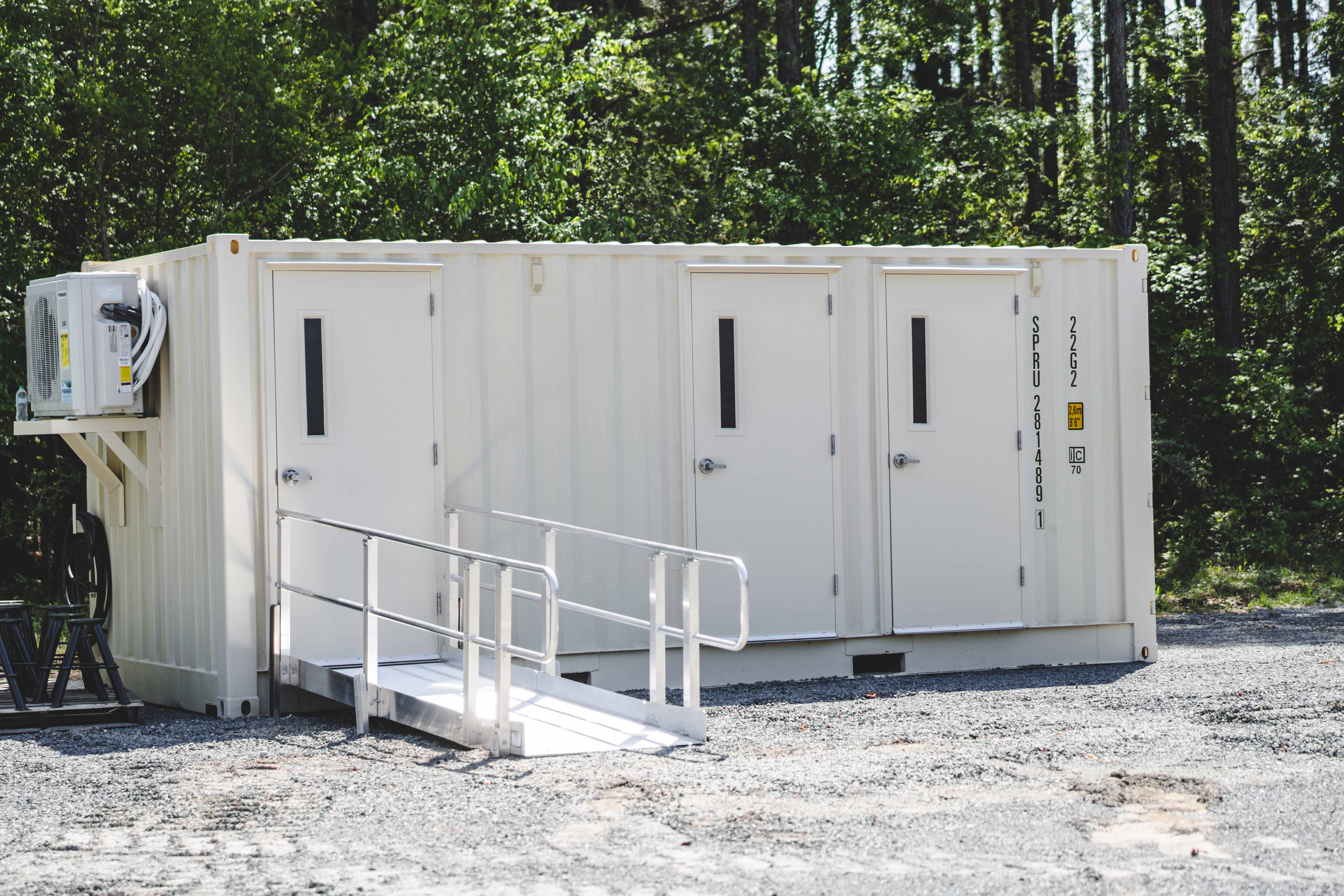 Exterior view of a white shipping container modified into a restroom facility with three doors and a handicap-accessible ramp. The unit is placed outdoors on a gravel lot surrounded by trees.