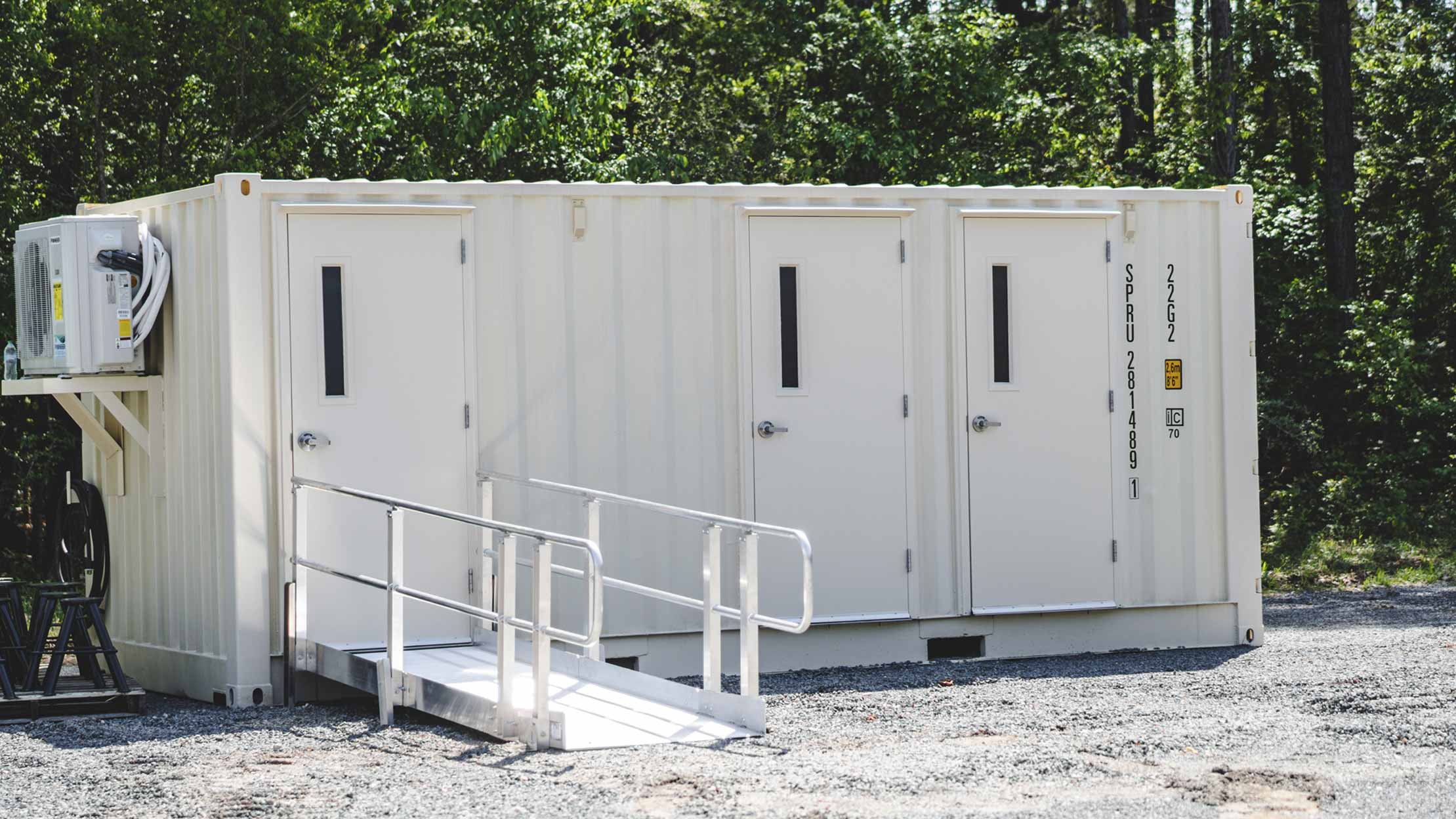 Exterior view of a white shipping container modified into a restroom facility with three doors and a handicap-accessible ramp. The unit is placed outdoors on a gravel lot surrounded by trees.