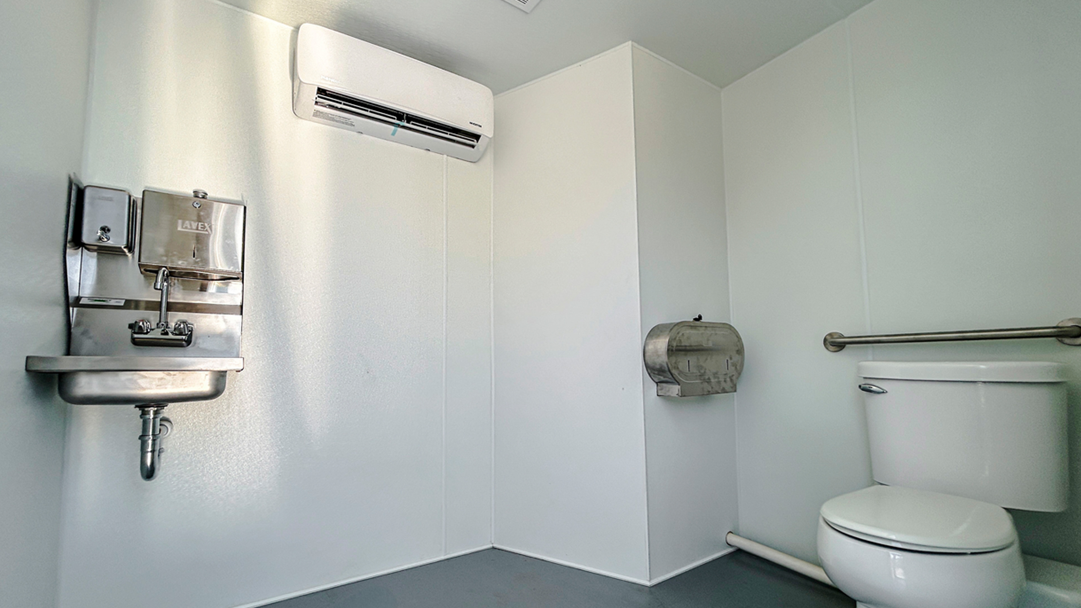Interior view of a modern, handicap-accessible restroom inside a white shipping container. The space features a stainless steel sink with a soap dispenser and paper towel dispenser, a white toilet, a metal toilet paper holder, and a wall-mounted mini-split air conditioning unit.