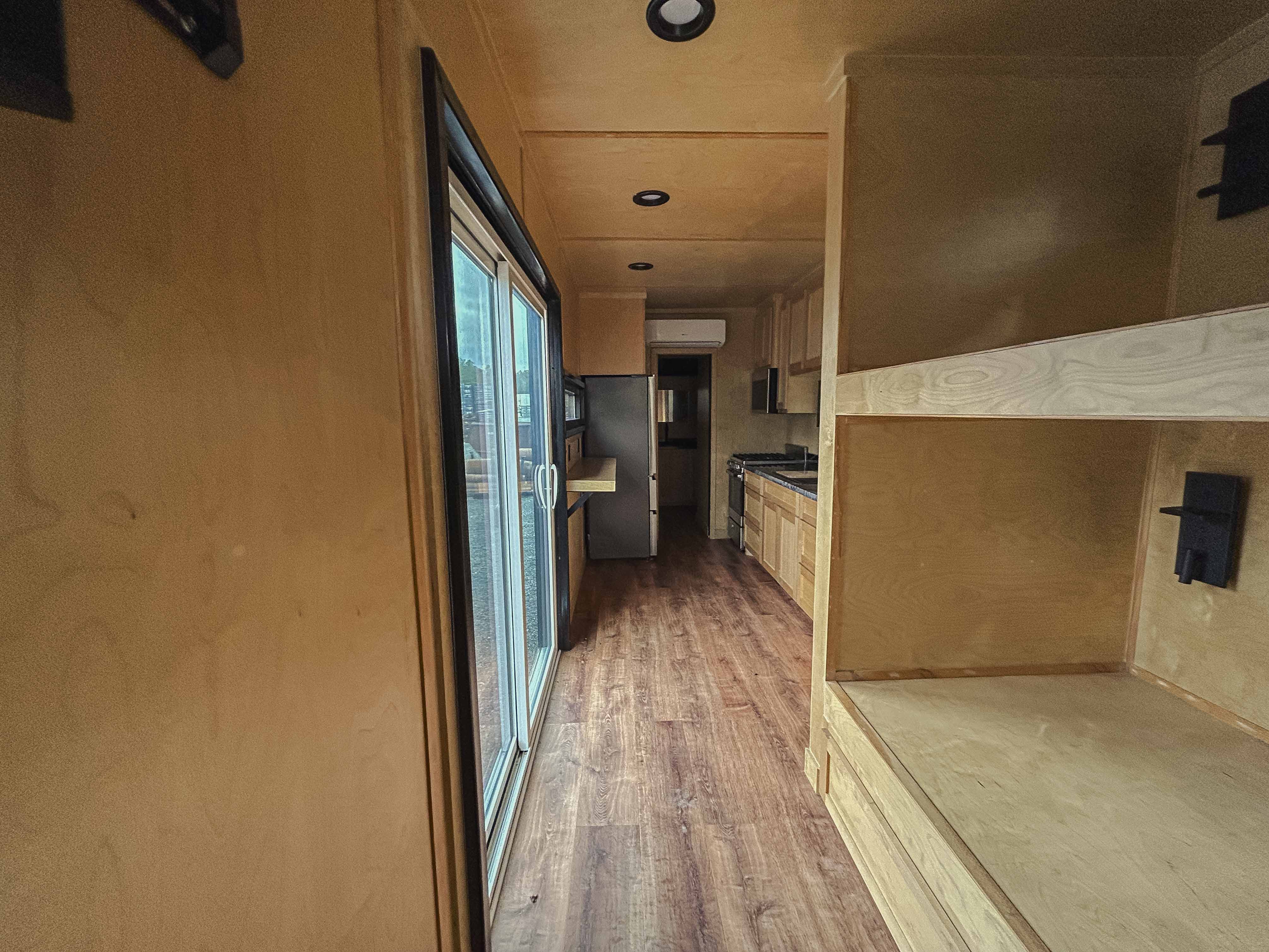 Interior hallway of a custom shipping container hunting cabin featuring built-in bunks, sliding glass door, recessed lights, and durable wood flooring—ideal for off-grid or camp housing.