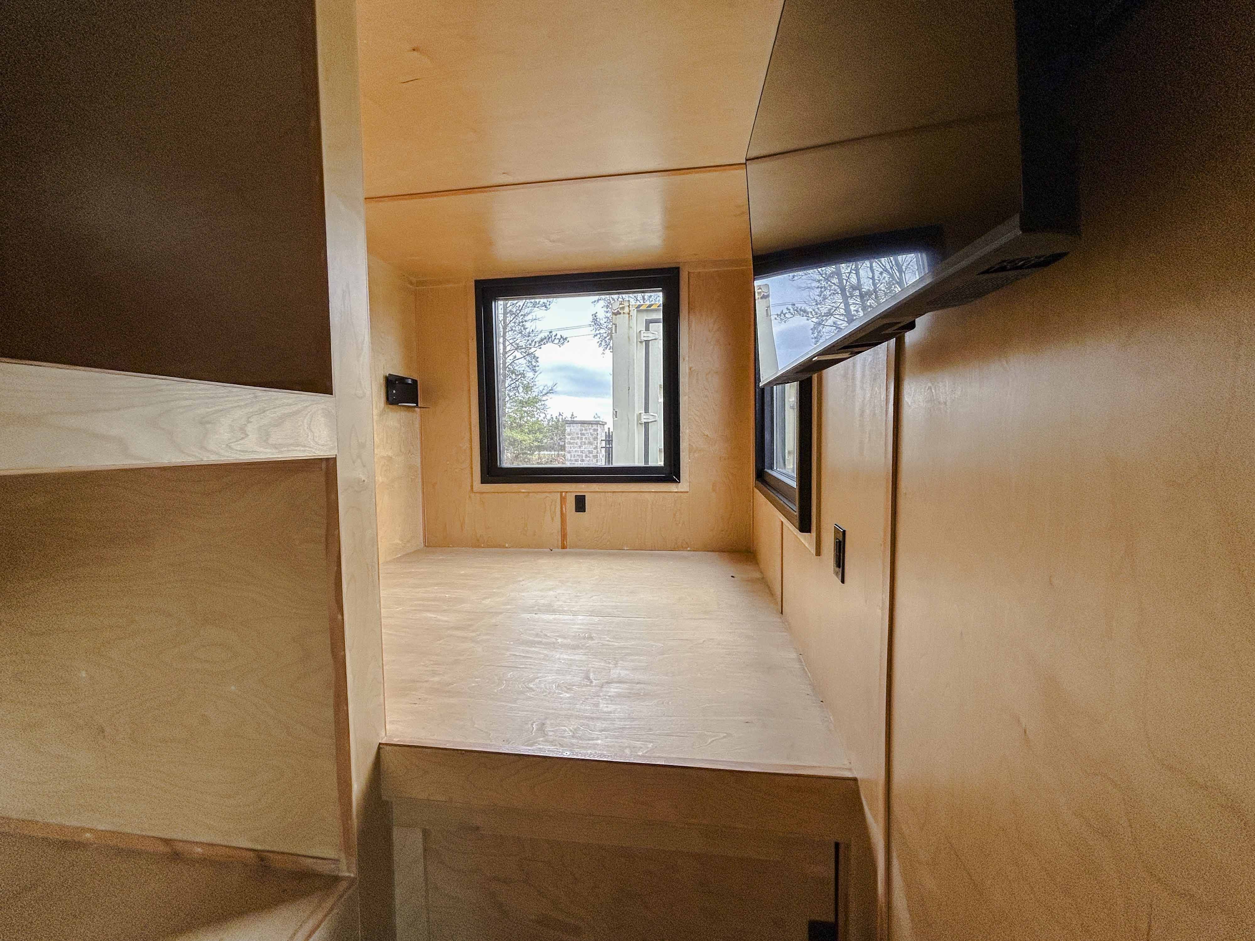 Custom shipping container hunting cabin interior featuring a built-in elevated bunk, natural wood walls, large modern windows, and mounted TV—compact sleeping area inside a converted container home.
