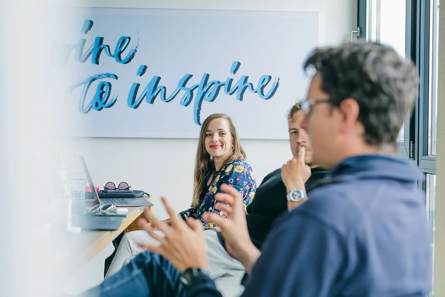 Drei Personen in einem Meetingraum, eine Frau lächelt, während zwei Männer aufmerksam zuhören.