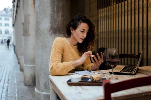 Publisher of a wine blog is checking her posts, while working outside of a bar,, with a glass of wine on the table.