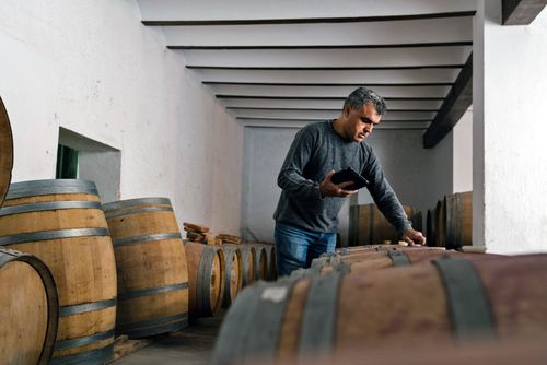 Producer inspects a barrel filled with an alcoholic beverage, and is noting his findings on an electronic tablet.