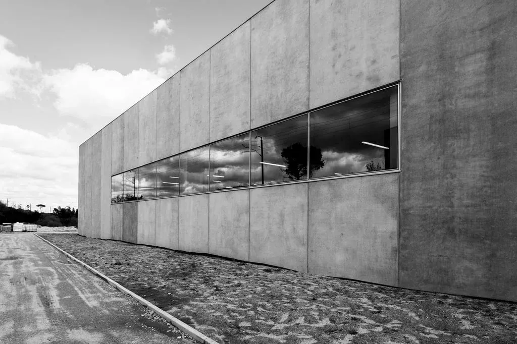 Fachada lateral de um prédio de concreto com janela longa refletindo nuvens e árvores.