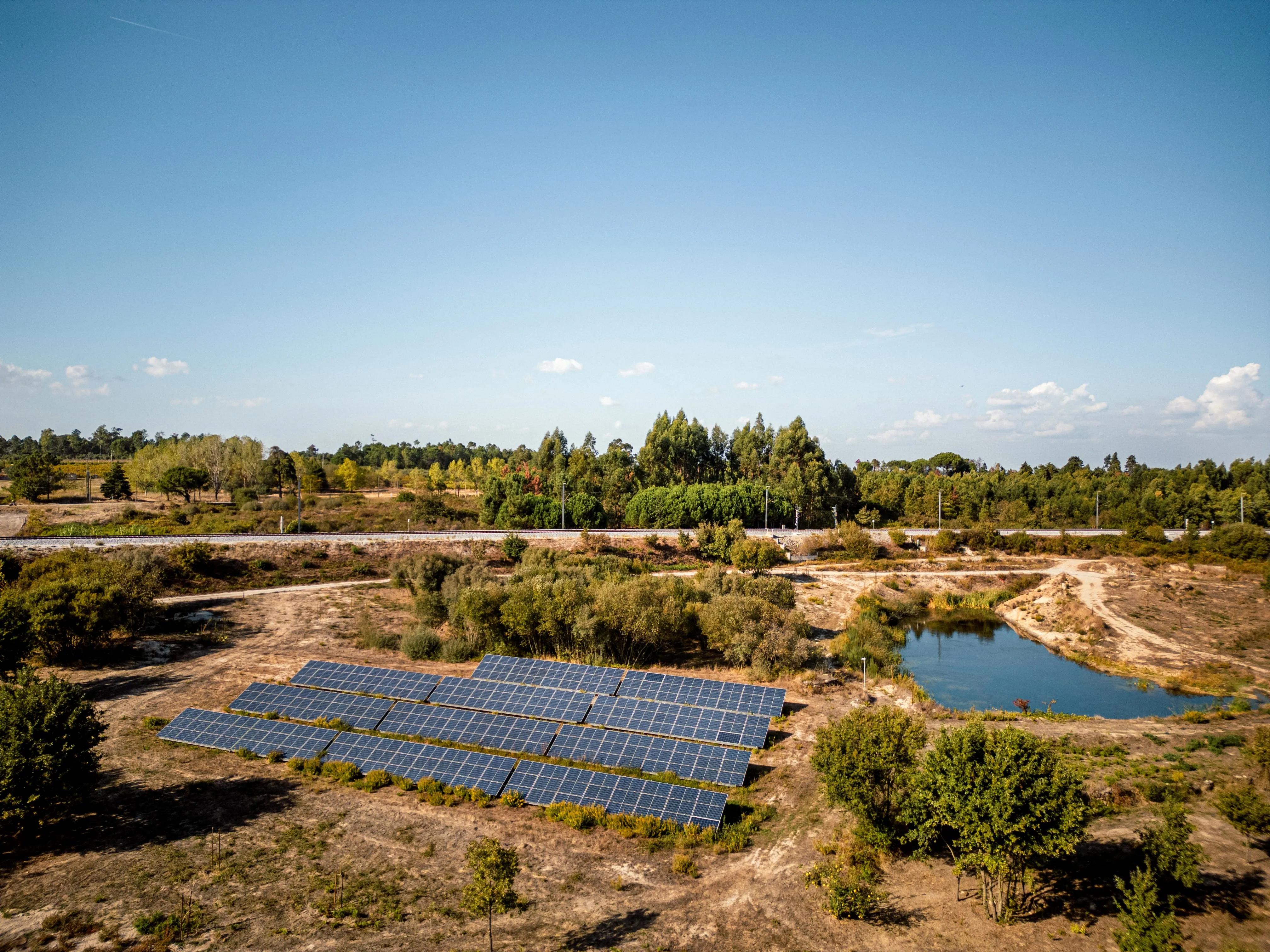 Painéis solares instalados em área seca próxima a um pequeno lago e árvores sob céu azul claro.