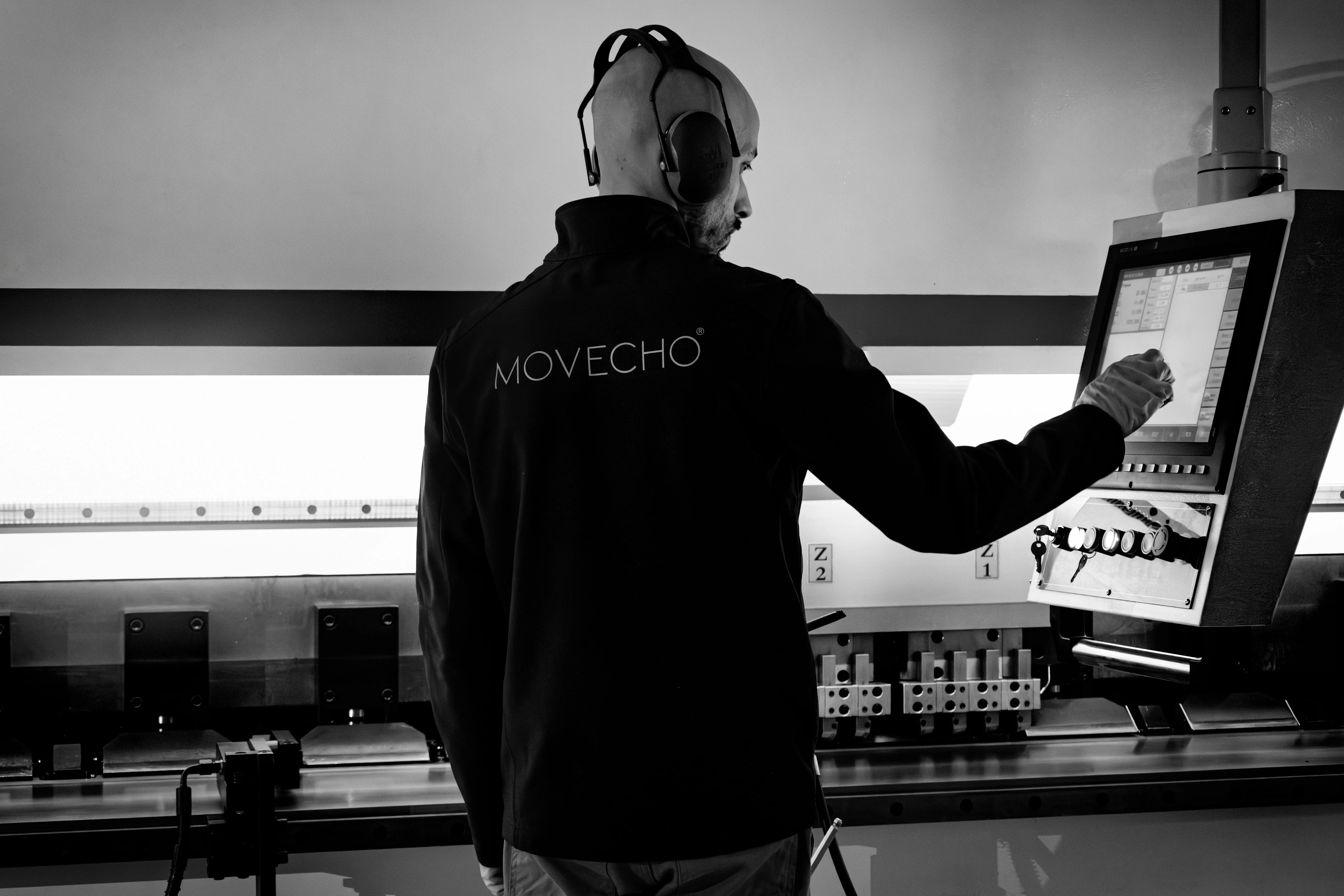 Operador em jaleco com logo MOVECHO usando protetor auditivo enquanto trabalha em máquina industrial.